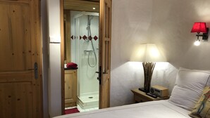 A bedroom with a bed, a nightstand with a lamp, a wooden door, and a shower area. - South-facing apt at the foot of the ski run. View of Tarentaise valley (Montvalezan)