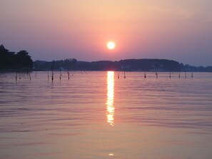 Un coucher de soleil sur une étendue d'eau calme, avec des bateaux et des arbres en arrière-plan.