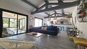 Un salon moderne avec un canapé, une table basse et un fauteuil en osier. On aperçoit à l’arrière-plan un coin cuisine avec des tabourets de bar et des luminaires suspendus.