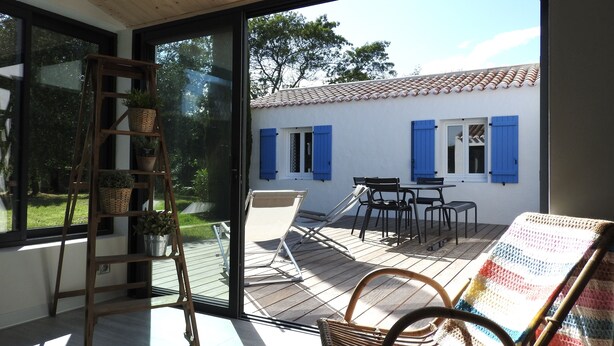 Une pièce dotée d'une porte-fenêtre coulissante donnant sur une terrasse extérieure avec une table et des chaises, un fauteuil en osier et un escabeau sur lequel sont disposées des plantes en pot.