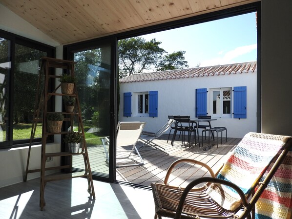 Habitación con puerta de vidrio corredera que da a un patio al aire libre con mesa y sillas, una silla de mimbre y una escalera con plantas en macetas.