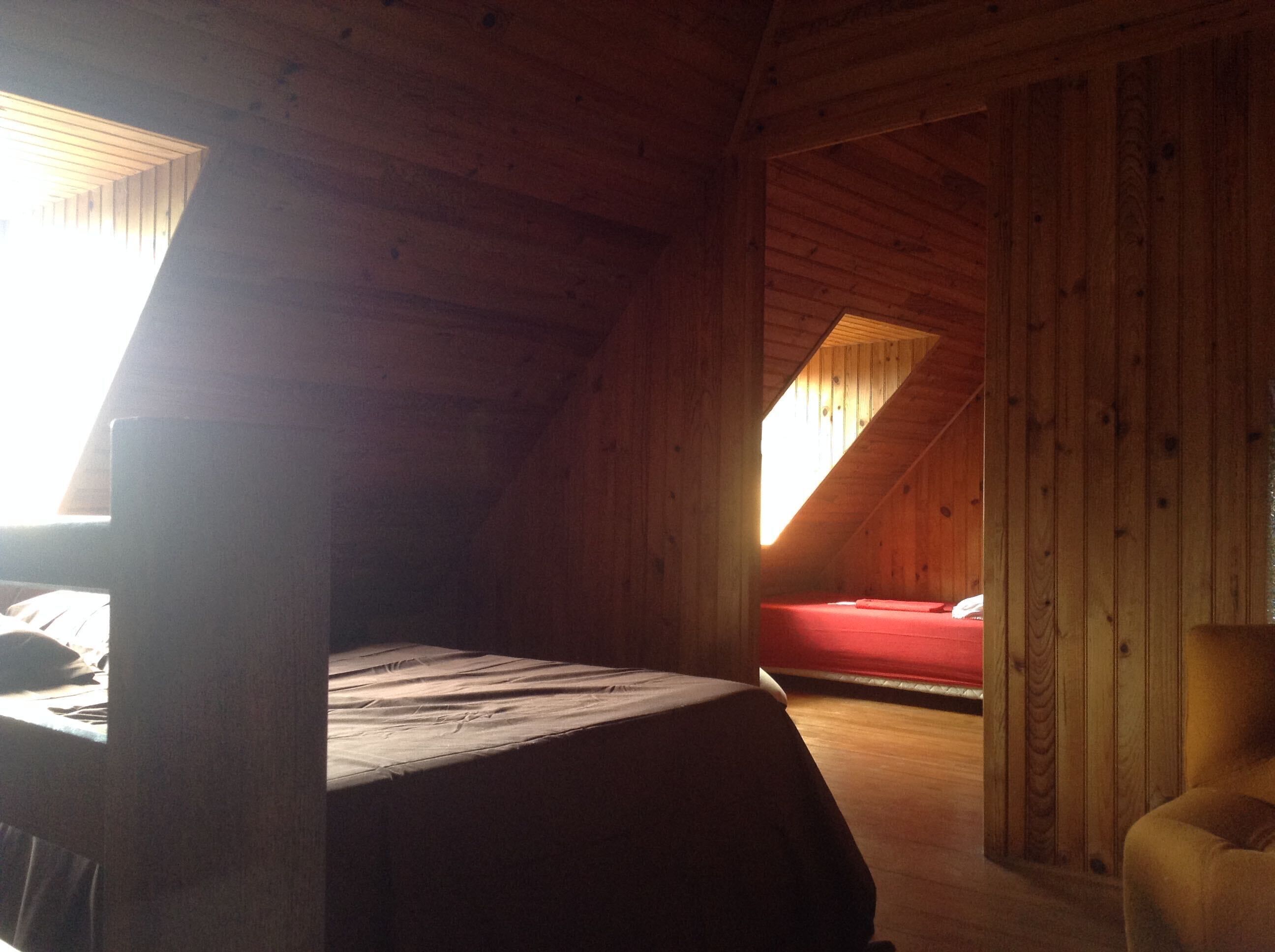 Une pièce avec un plafond en pente, des murs en bois, un lit recouvert d’une couverture rouge et une chaise.