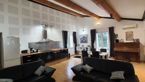 A modern living room with a kitchen area, featuring a large black sofa, a dining table, and a wooden ceiling.