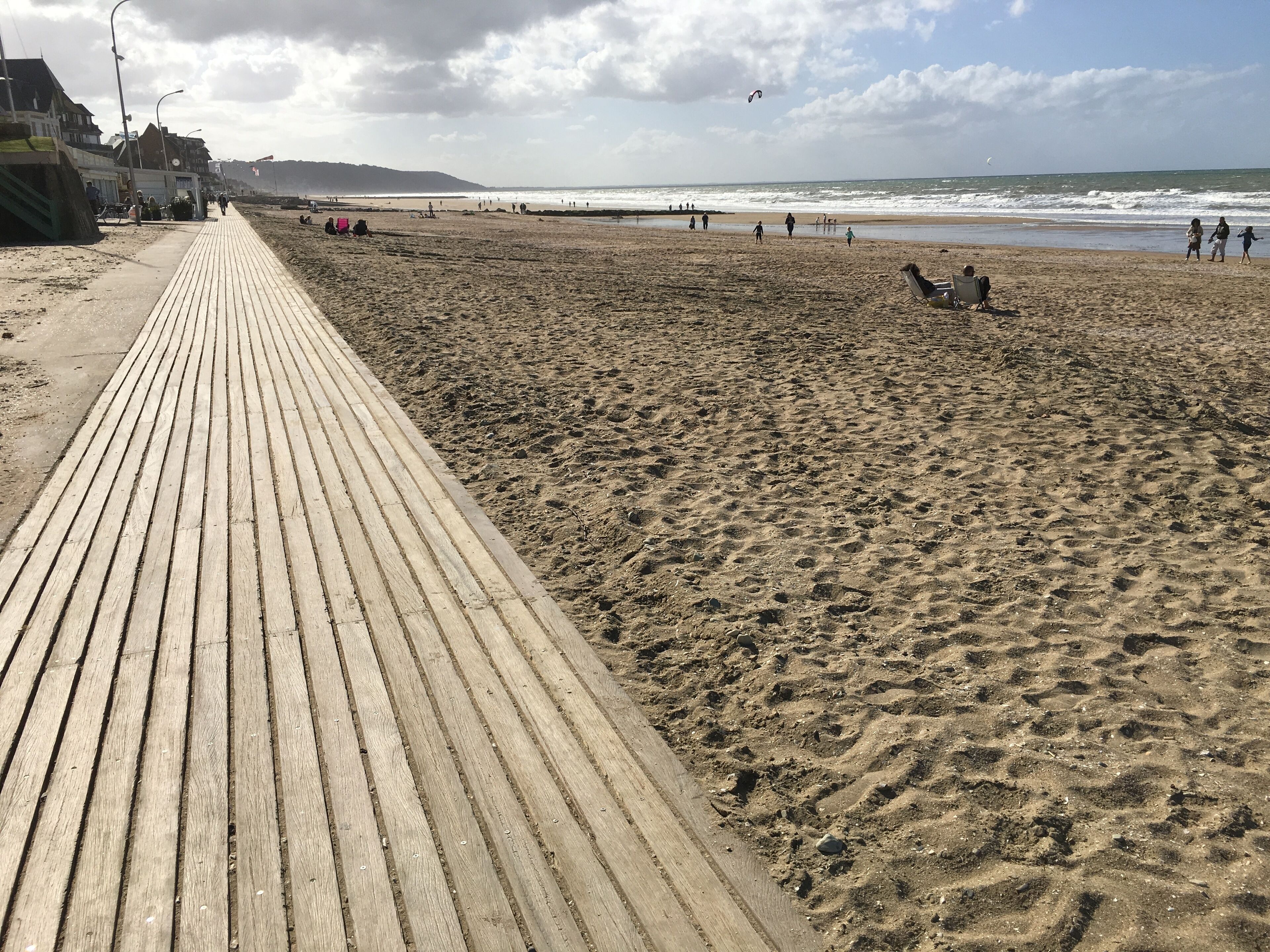 Ein hölzerner Steg führt zu einem Sandstrand mit Blick auf das Meer und sanfte Hügel in der Ferne.