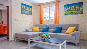 A living room with a grey sofa, yellow and blue cushions, a glass coffee table with a flower arrangement, and two paintings on the walls.