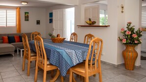 A dining area with a table set for six, surrounded by wooden chairs. A fruit bowl is on the table, and a potted plant is on the right.