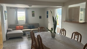 A living room with a sofa, a dining table with chairs, a potted plant, and a window with curtains. - VILLA LODGE WITH ROOFTOP JACUZZI (sainte-anne)