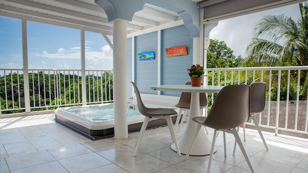 A covered outdoor area with a hot tub, a table with chairs, and a view of the surrounding greenery.