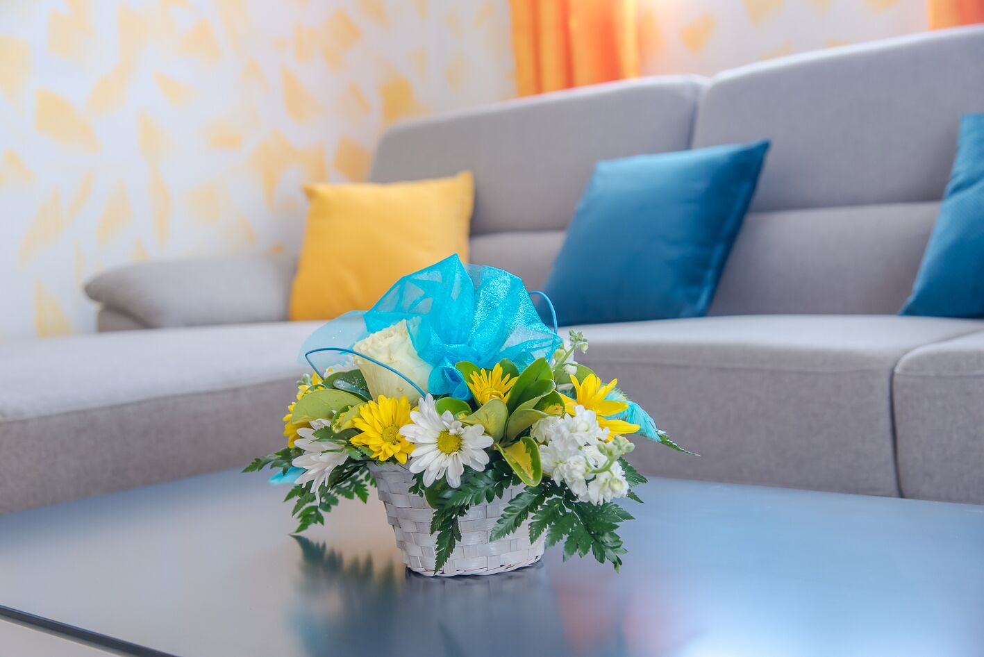 Een boeket bloemen op een tafel in een woonkamer met een grijze bank en blauwe kussens.