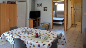 A room with a dining table covered in a tablecloth with a pattern, a television on a stand, a bed, and a shower area. - Single-storey furnished gite in the heart of the village (Bellefontaine)