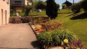 A paved outdoor area with a seating arrangement, a garden with various plants, and a building in the background. - Single-storey furnished gite in the heart of the village (Bellefontaine)