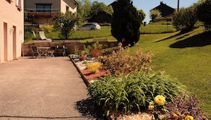 A paved outdoor area with a seating arrangement, a garden with various plants, and a building in the background. - Single-storey furnished gite in the heart of the village (Bellefontaine)