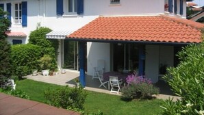 Une maison à deux étages avec un toit recouvert de tuiles rouges, un patio couvert avec des meubles d’extérieur et un jardin bien entretenu.