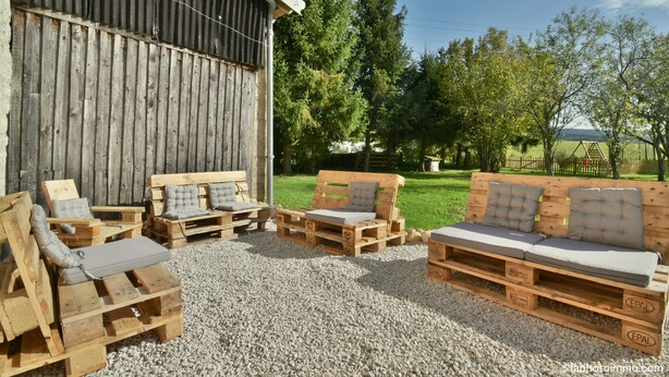 Coin salon extérieur aménagé avec des palettes en bois et des coussins, installé sur un sol de gravier dans un cadre rural, avec une grange et des arbres en arrière-plan.