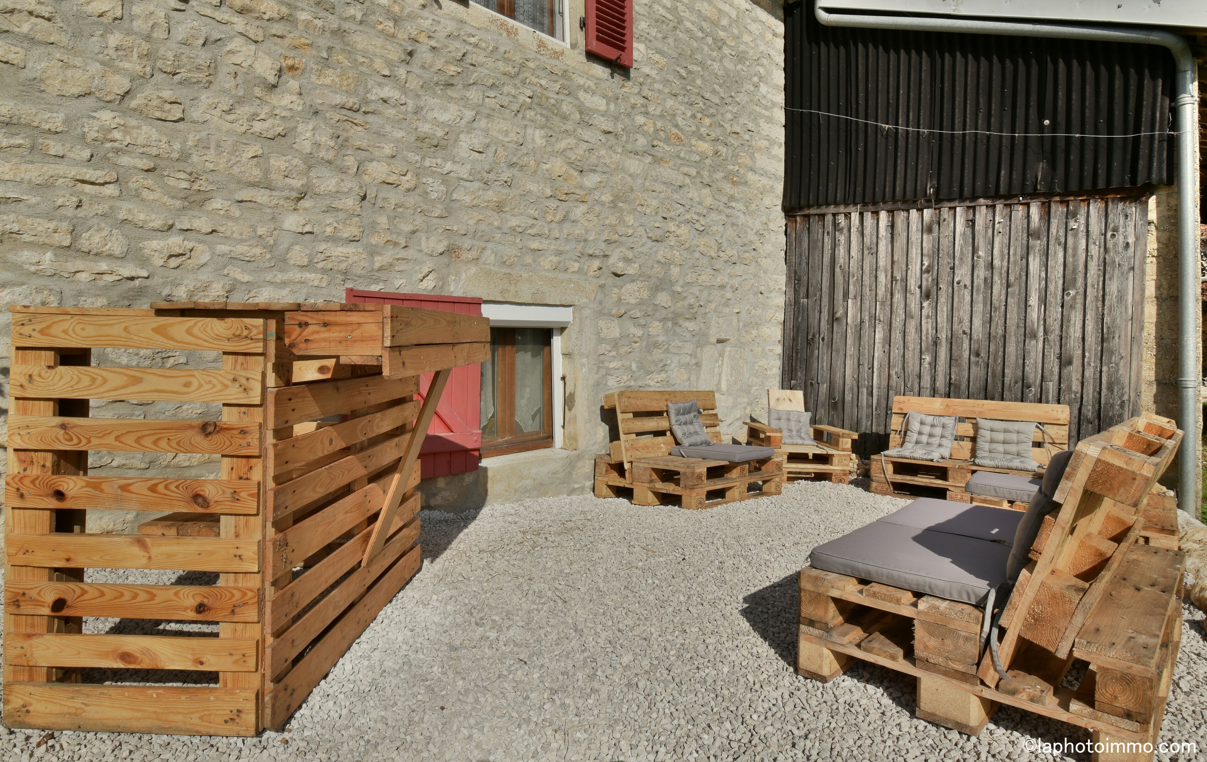 Zona esterna rustica con sedute in legno ricavate da bancali, affacciata su un muro in pietra e una recinzione in lamiera ondulata.