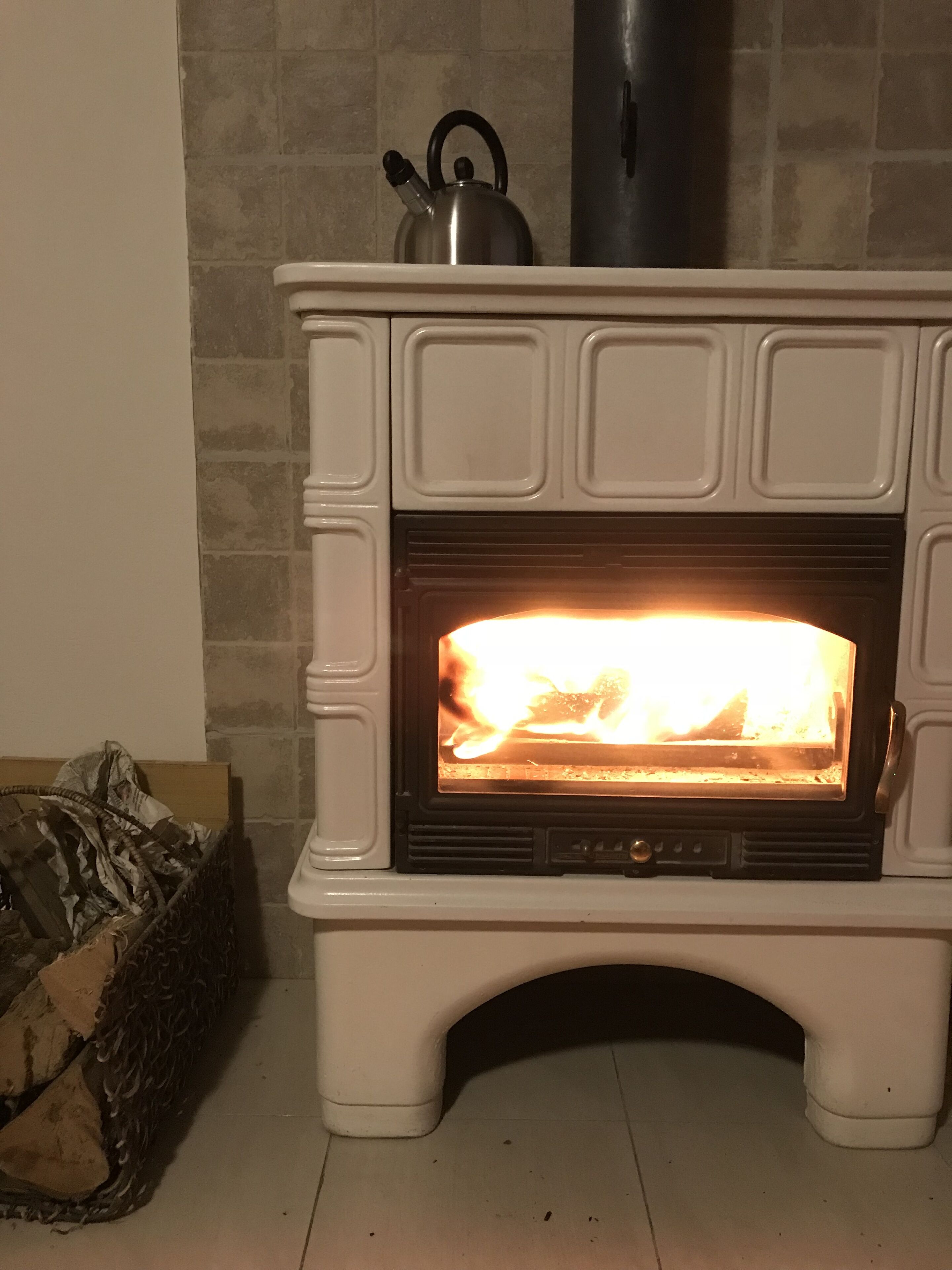 En vit vedeldad kamin med synliga lågor, en vattenkokare ovanpå och en förvaringslåda för ved till vänster.