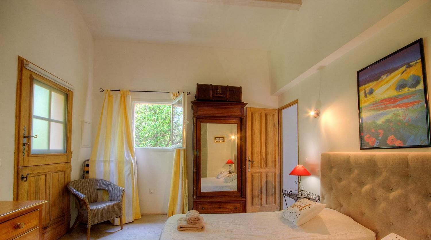 Une chambre à coucher comprenant un lit, une chaise, une armoire, un miroir, un tableau et une fenêtre avec des rideaux.