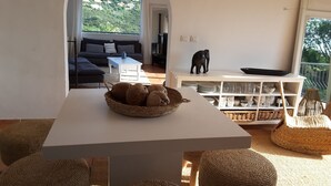 Un saló modern amb una taula blanca, uns poufs teixits, un cistell de pa i una vista d'una vessant de turons verda a través d'una finestra allargada.