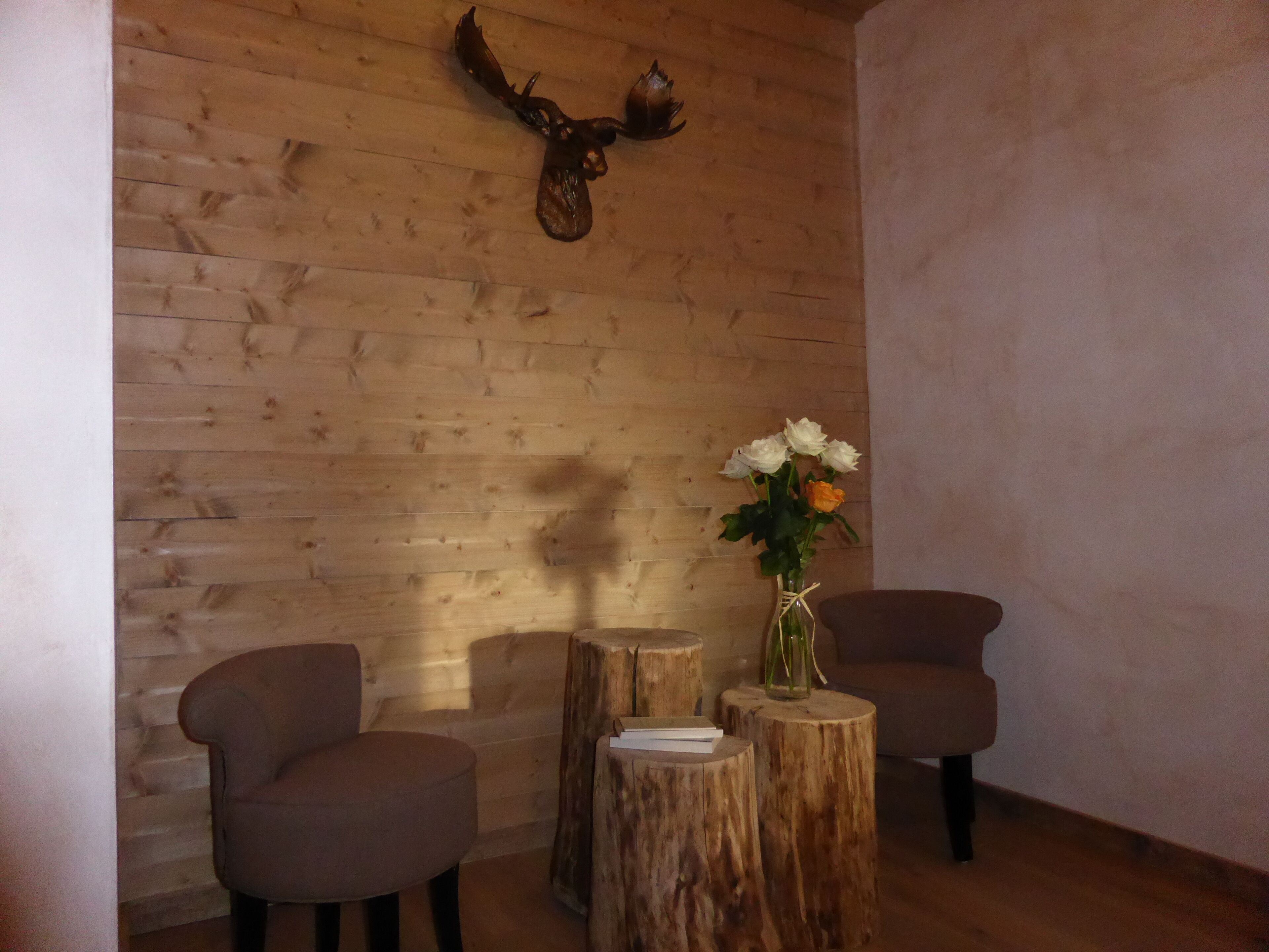 Une pièce aux murs en bois, ornée d’une tête de cerf, avec deux chaises et un vase de fleurs posé sur un tabouret en bois.