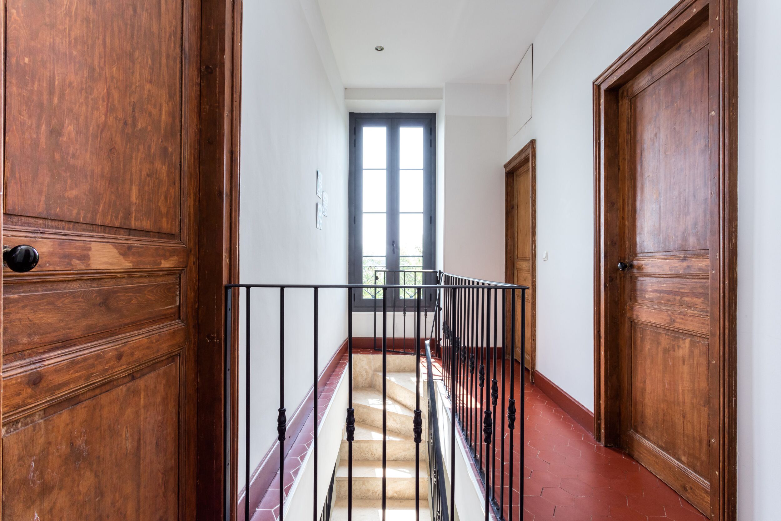 A hallway with wooden doors, a black metal railing, and a red-tiled floor.