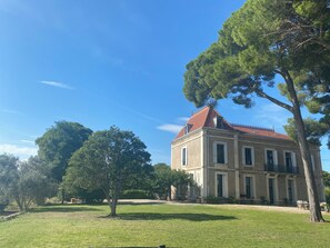 Un grand bĂątiment historique, dotĂ© dâun toit recouvert de tuiles rouges, entourĂ© dâune pelouse bien entretenue et de plusieurs arbres.