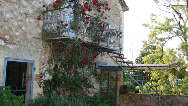 Un bâtiment rustique en pierre, doté d’un balcon orné de fleurs rouges, d’une porte en bois et d’un escalier menant à un étage supérieur.