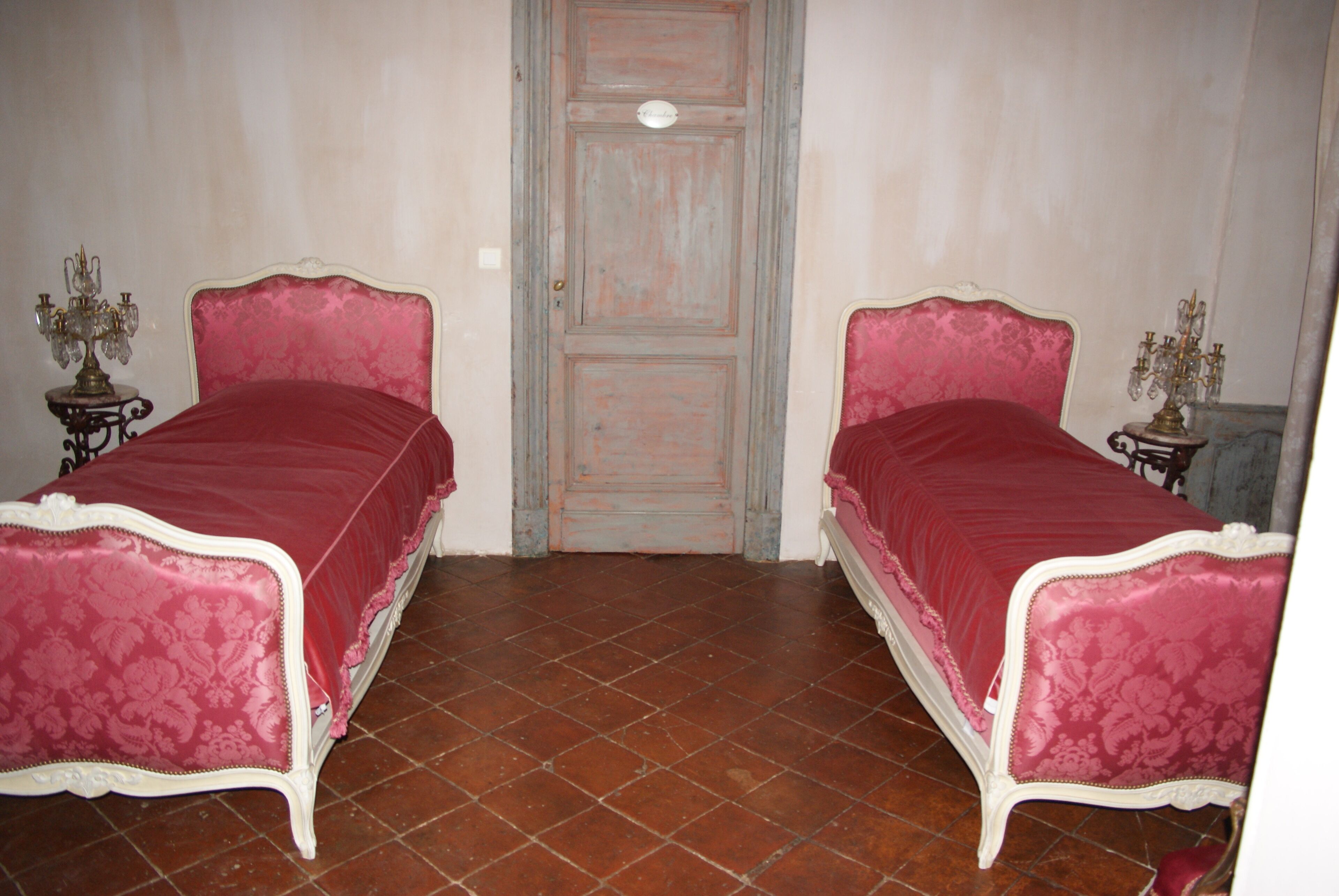 A room with two beds, each with a red bedspread and white trim, a wooden door, and a tiled floor.