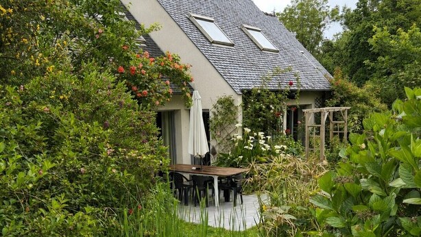 A house with a sloped roof, a patio with a table and chairs, and a garden with various plants and flowers. - Peaceful house between sea and forest near Perros-Guirec (Louannec)