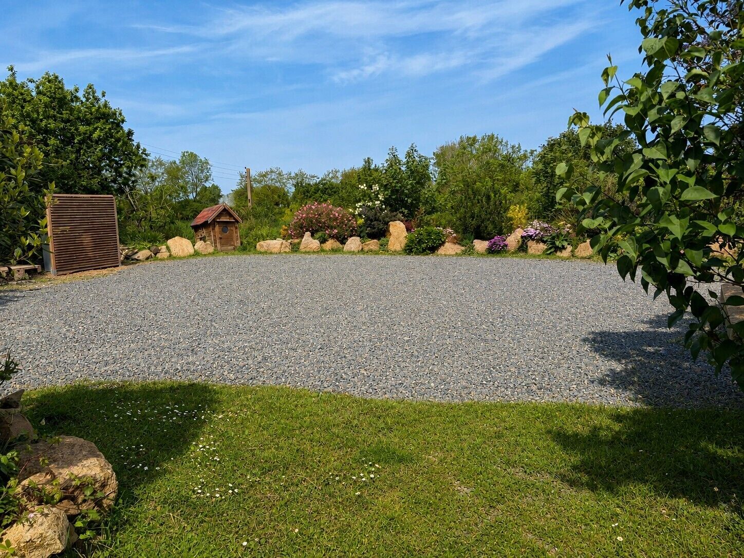A gravel parking area with a small wooden structure, surrounded by greenery and rocks.