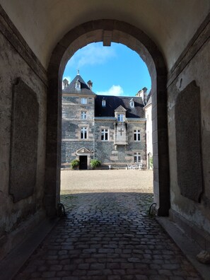 Un couloir en arc de cercle, avec un chemin pavé menant à un bâtiment doté d’une façade en pierre et de nombreuses fenêtres.
