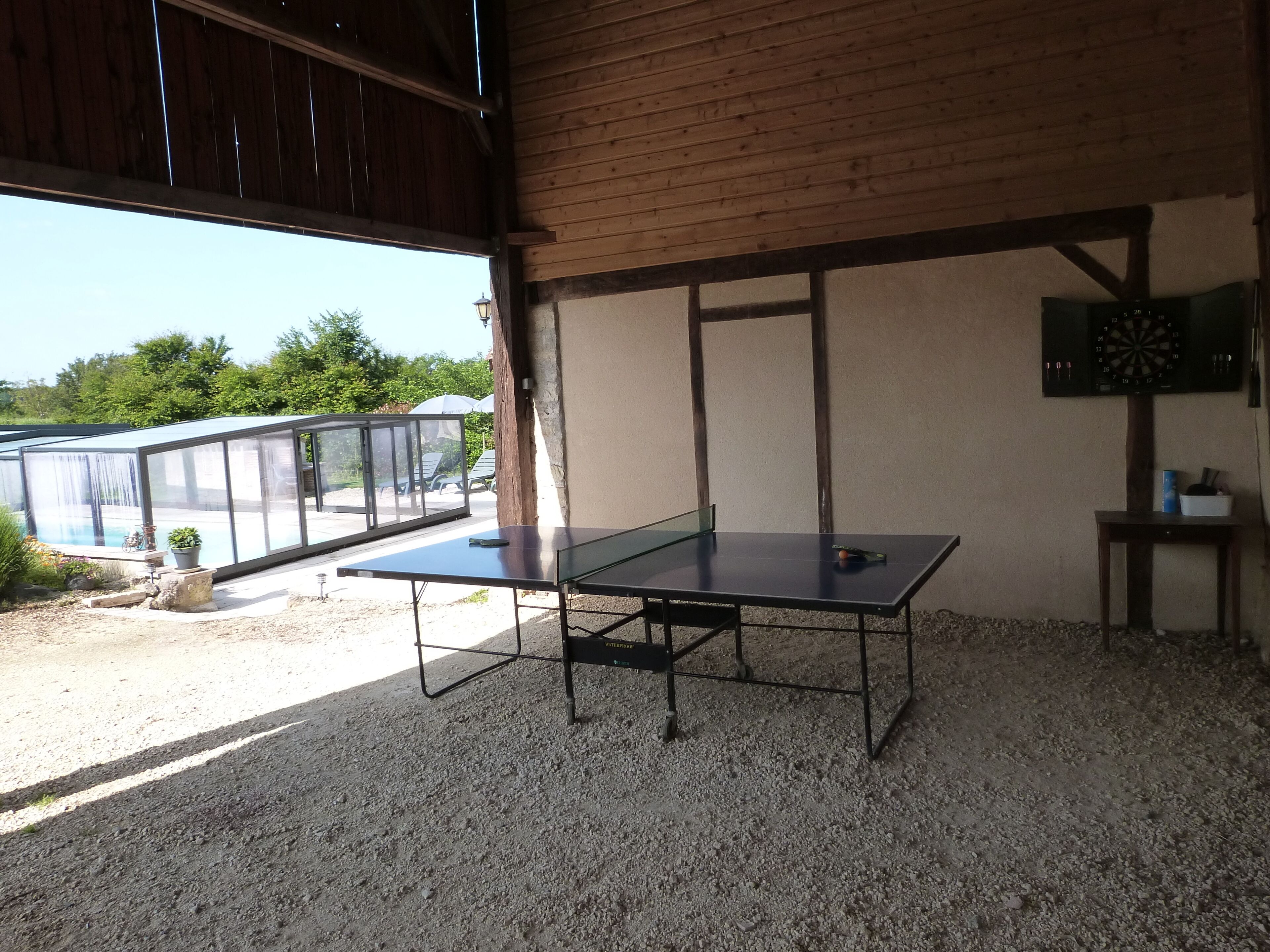 A ping pong table in a covered outdoor area with a wooden ceiling and a dartboard on the wall.