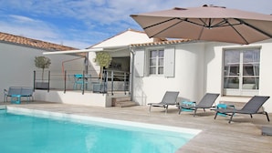 Un espace aménagé au bord de la piscine offrant une vue dégagée sur celle-ci, un patio couvert avec des chaises et une table, et un bâtiment doté d’un toit en tuiles et de fenêtres.