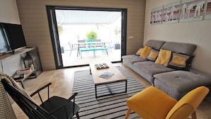 Un salon avec un canapé gris, des coussins jaunes, un fauteuil noir, une table basse et un téléviseur posé sur un meuble.