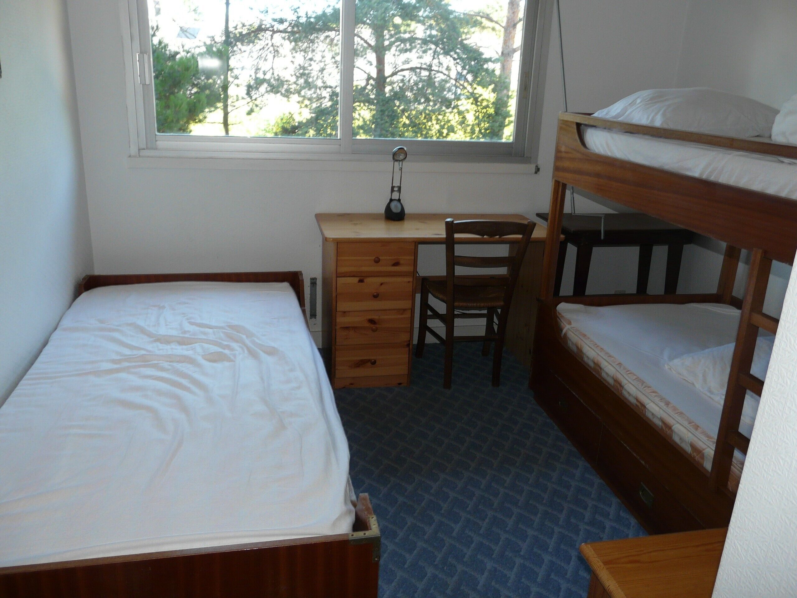 Ein Schlafsaalzimmer mit Etagenbett, einem Einzelbett, einem Schreibtisch mit Stuhl und einem Fenster mit Blick auf Bäume.