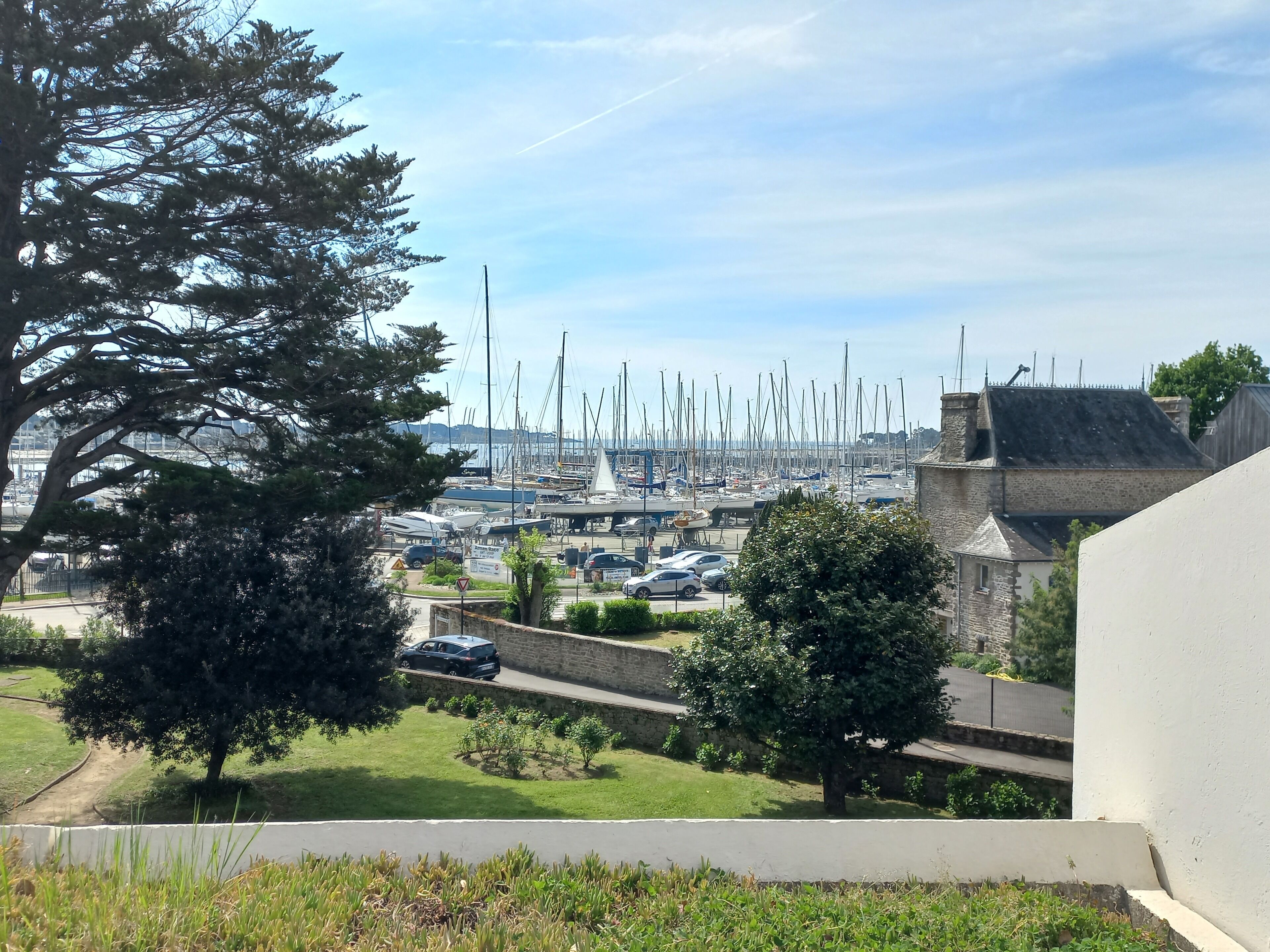 A marina with numerous boats docked, a parking area with cars, and a stone building with a sloped roof.