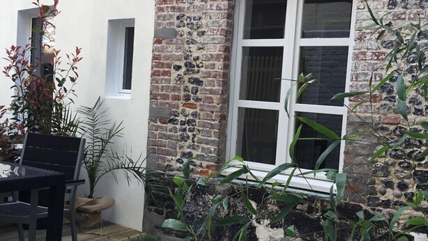 Il s’agit d’un bâtiment rustique, avec une façade en briques et un mur blanc. On y aperçoit une fenêtre à carreaux et une porte. Une terrasse en bois est visible devant le bâtiment.