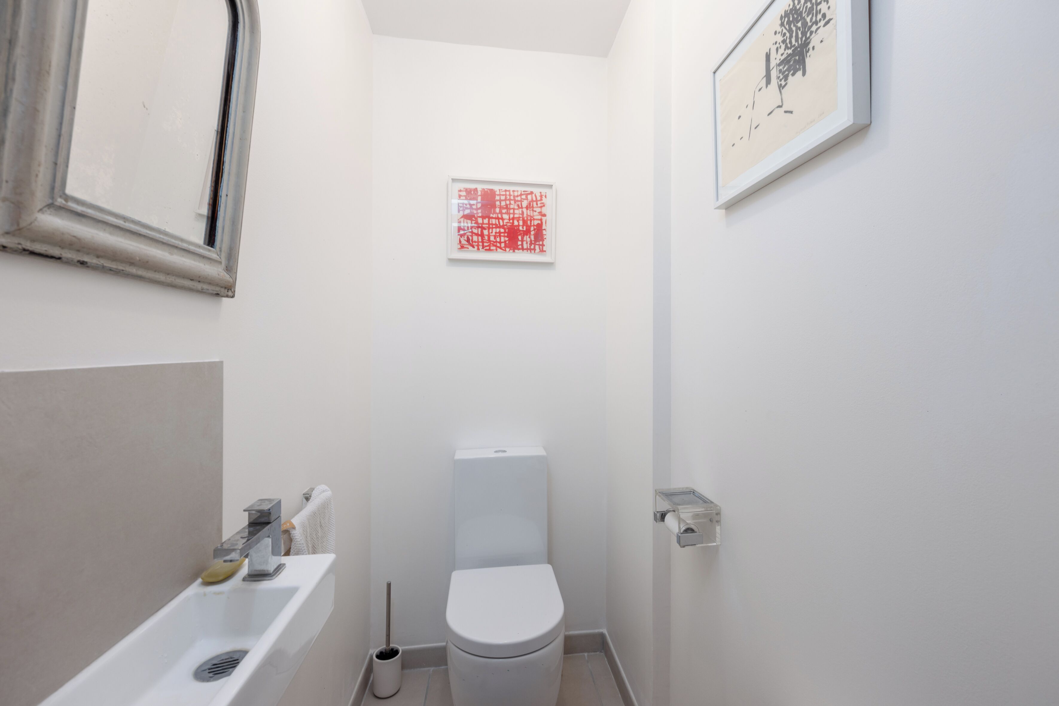 A small, white bathroom with a toilet, a sink, and a mirror. There are two framed pictures on the walls.