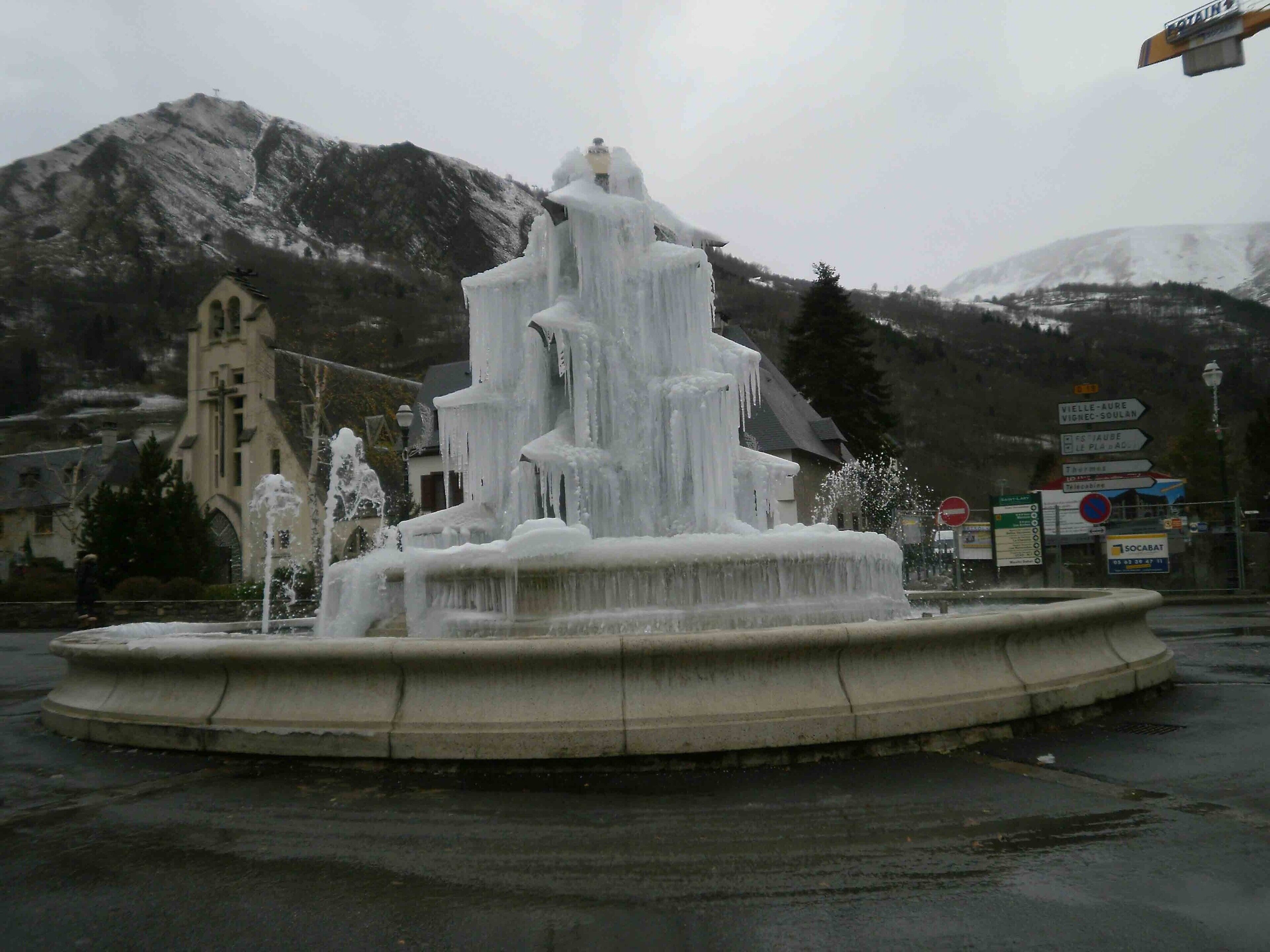 A multi-tiered fountain with water jets, surrounded by a circular stone basin, set against a backdrop of a church and mountains.
