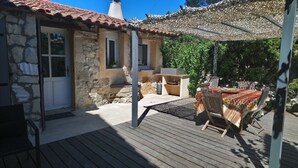Un edificio de piedra con techo de tejas, terraza de madera y zona de estar al aire libre cubierta, con una mesa y sillas.
