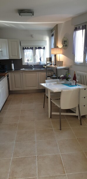 Een moderne keuken met witte kastjes, een eettafel met stoelen, een raam met gordijnen, en een lamp op het aanrecht.