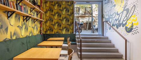 A cafe with wooden tables and green cushioned seating, a staircase leading to an outdoor area, and a mural on the wall.