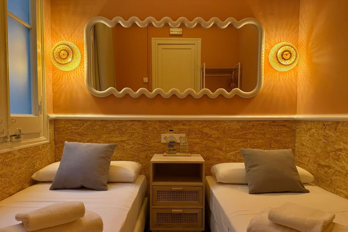 A hotel room with two single beds, a large mirror with a wavy frame, and a small table with a bottle and glass.