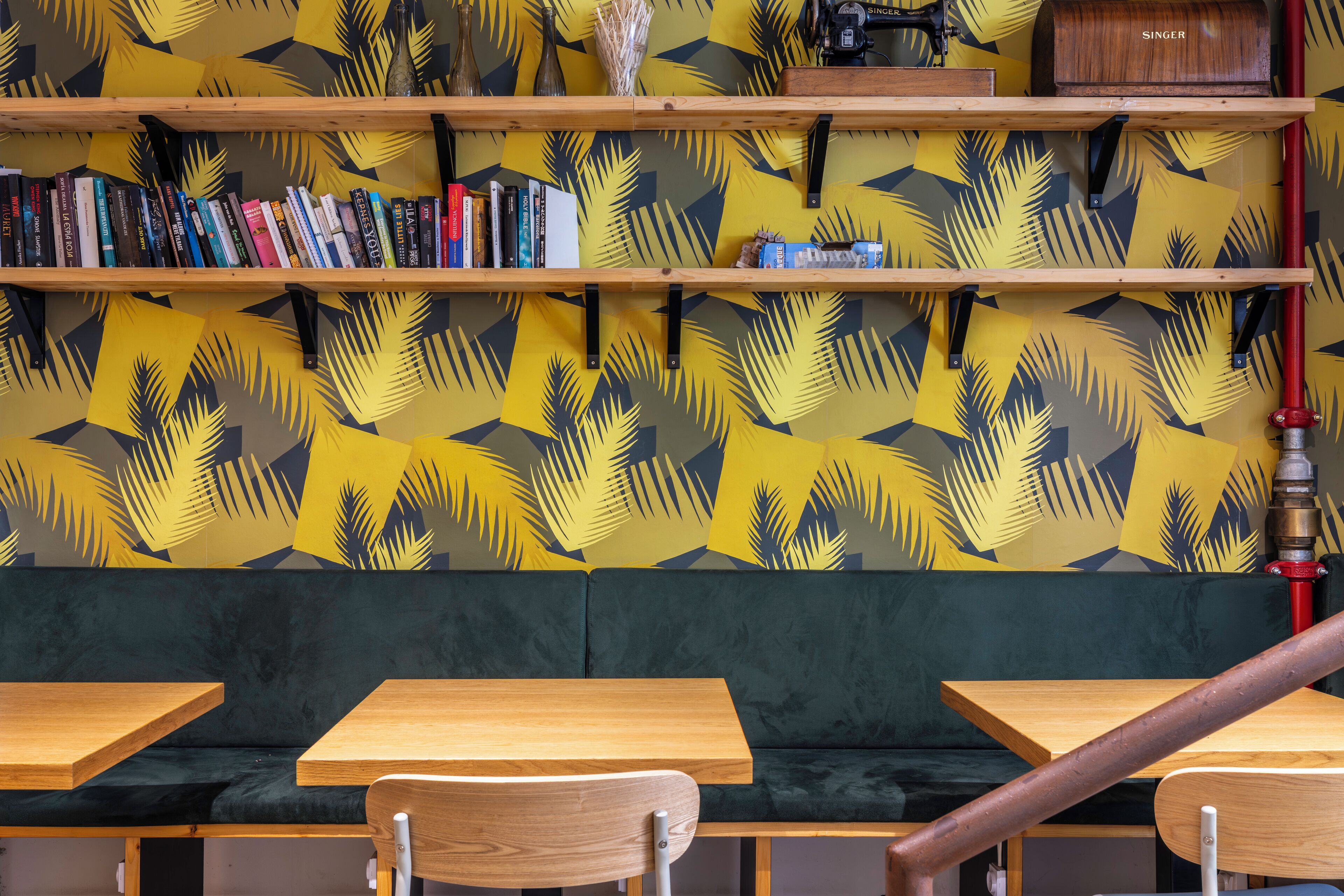 Eine Sitzecke mit Holztischen und -stühlen, einer gepolsterten grünen Bank und Regalen mit Büchern und Dekorationen an einer Wand mit Muster.
