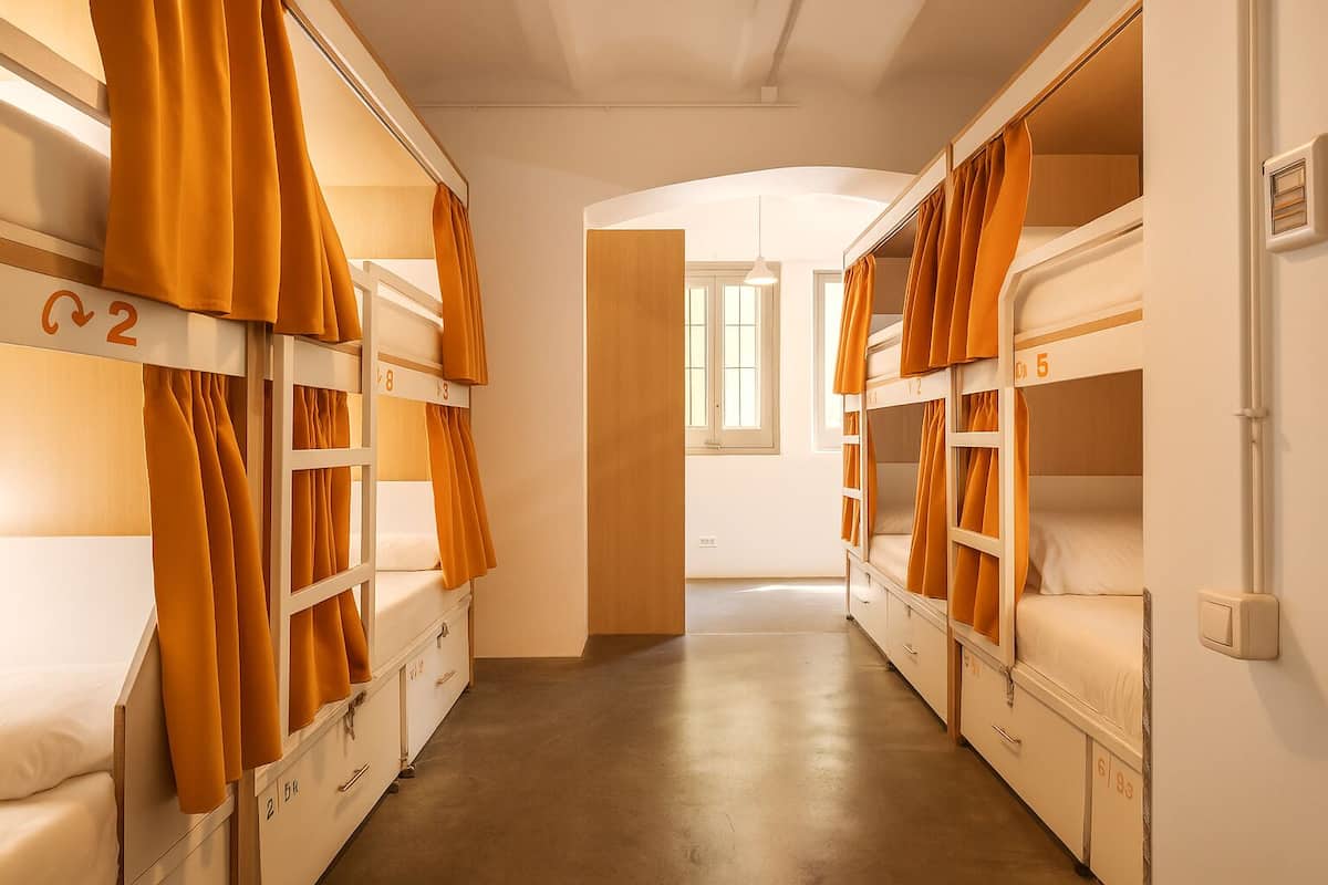A dormitory room with bunk beds, numbered beds, and a corridor leading to another room.