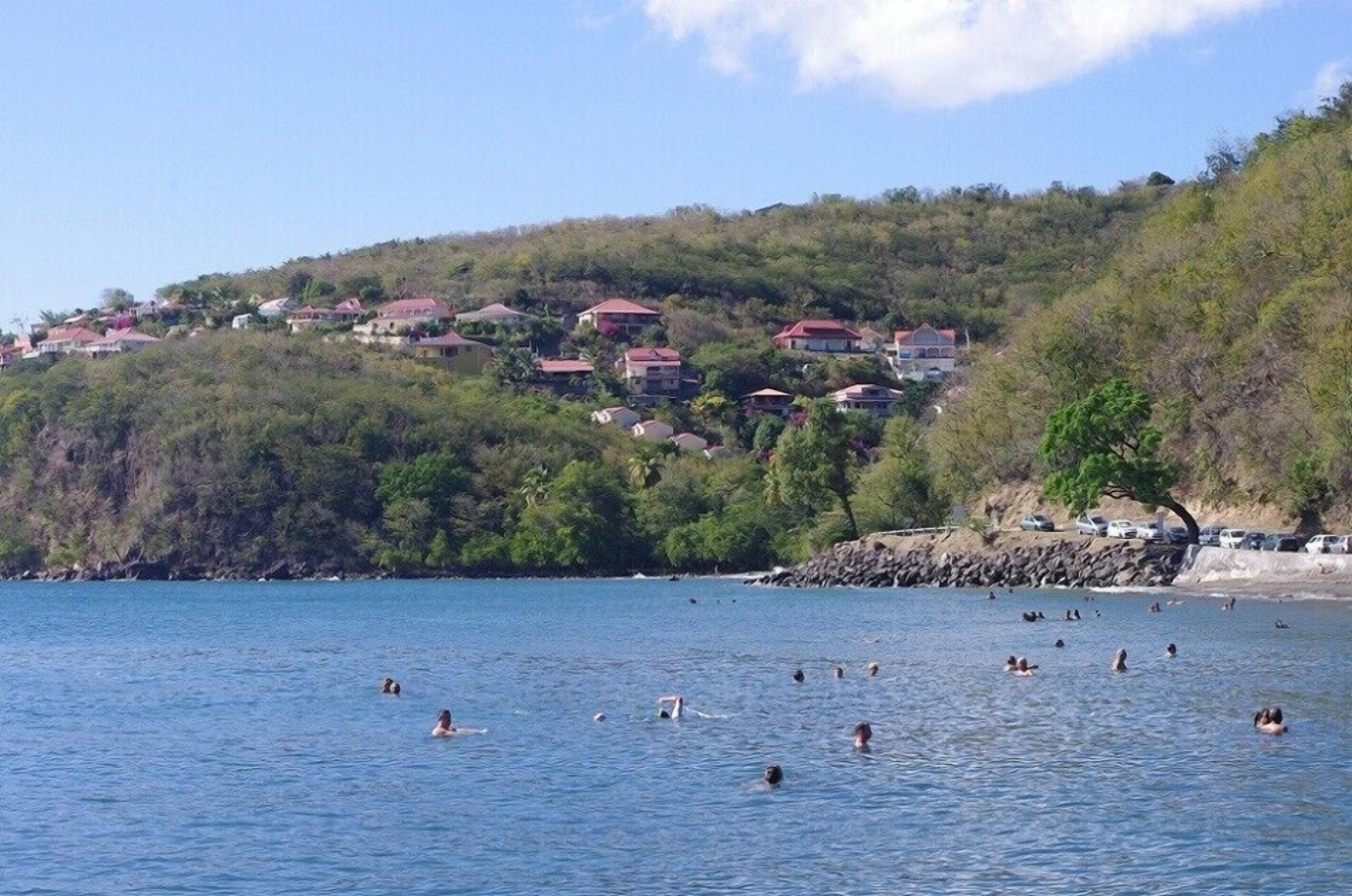 Zona costera con casas en una colina y gente nadando en el agua.