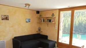 Ein Wohnzimmer mit einem schwarzen Ecksofa, einem kleinen Tisch mit Lampe, einem Regal mit Dekoration und einer großen Schiebeglastür mit Blick auf einen See und Wald.