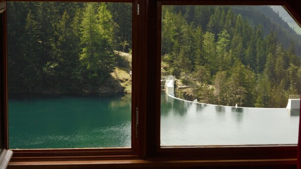 Eine Hütte mit einem großen Fenster mit Blick auf einen türkisblauen See und ein bewaldetes Gebiet.