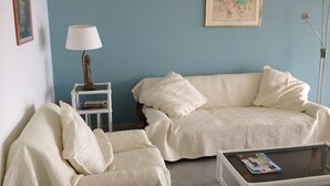 Een woonkamer met een blauwe wand, een witte bank, een witte fauteuil, een klein wit bijzettafeltje met een lamp, en een glazen salontafel.