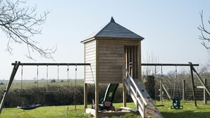 A wooden playhouse with a ramp and swings in a grassy area.