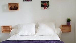 Una cama bien hecha con ropa blanca y una manta morada. Hay dos mesitas de noche de madera con pequeñas macetas.